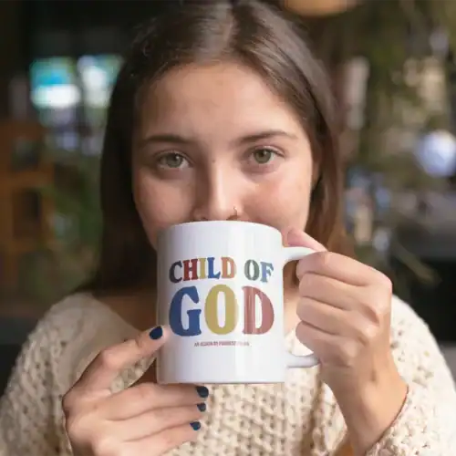 Colorful "Child of God" mug with bold text and an album reference, held by a young woman with brown hair in a cozy setting.