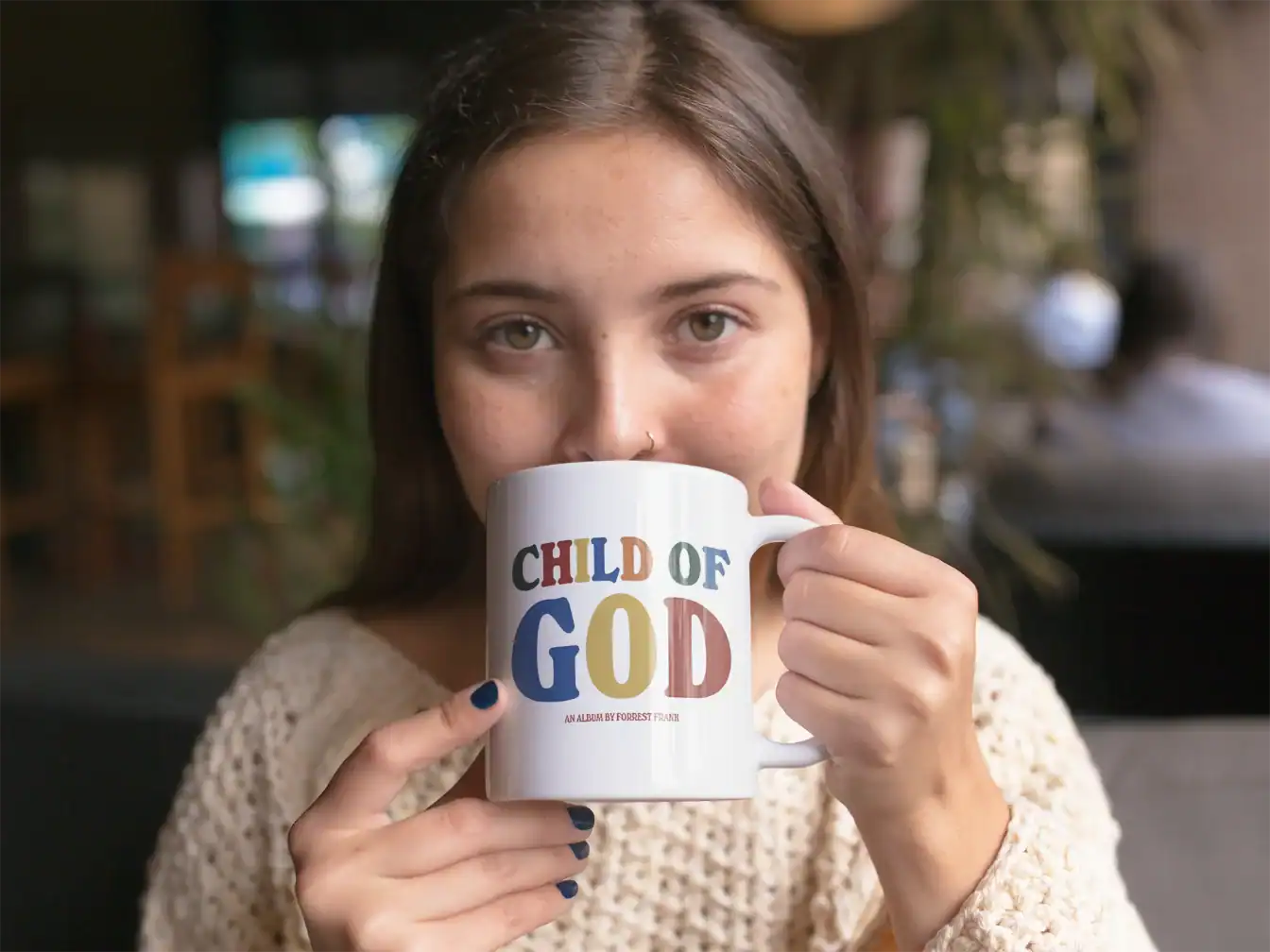 Colorful "Child of God" mug with bold text and an album reference, held by a young woman with brown hair in a cozy setting.