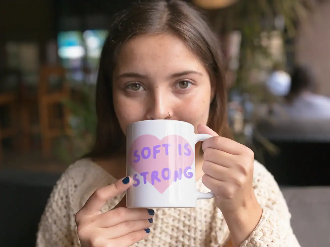 Cute young woman holding a white mug with the phrase "Soft is Strong" written on it in colorful letters.